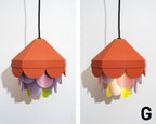 A sculptural pendant lamp with a terracotta orange top shade and layered pastel petal-like shades in pink, purple, yellow, and green, hanging against a white wall. A black cord suspends the lamp, and a black letter 'G' appears in the bottom right corner.